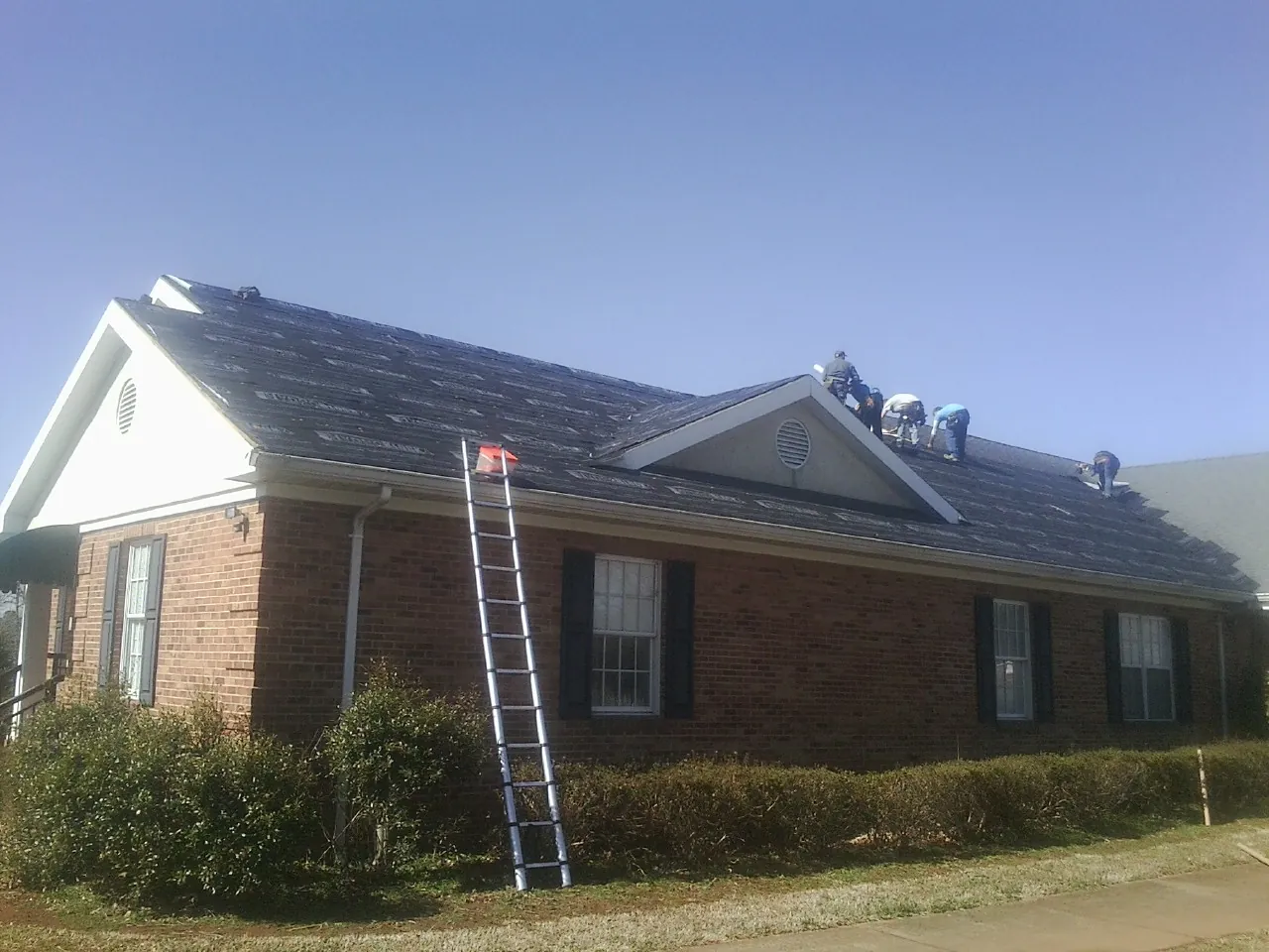 Crew tearing shingles off a brick ranch roof with a ladder set — Emergency Roof Repair work by our crew in Bovey