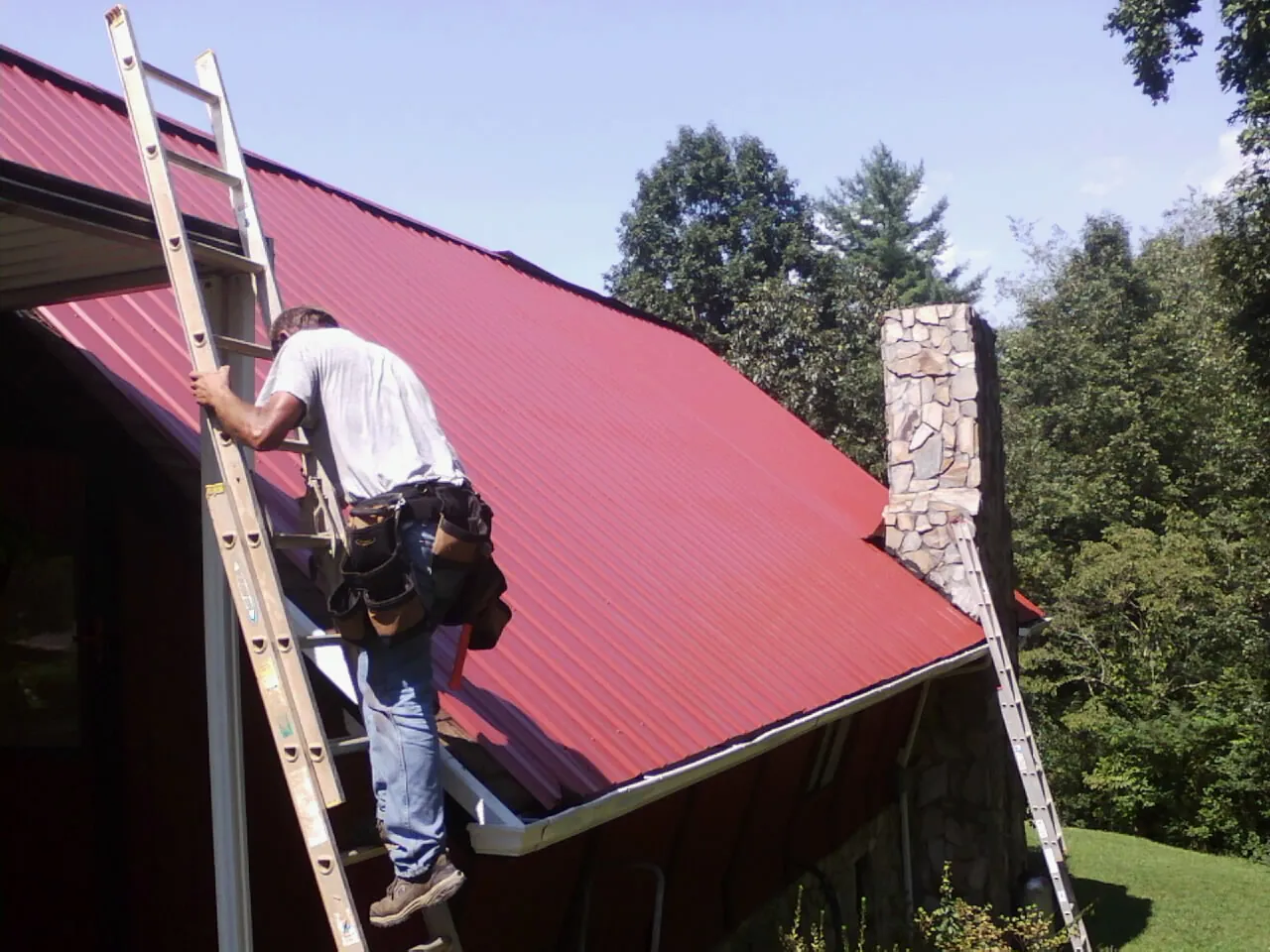 Roofer on a ladder beside a red metal roof and stone chimney — Soffit & Fascia Repair by Roofing Brothers in Bovey
